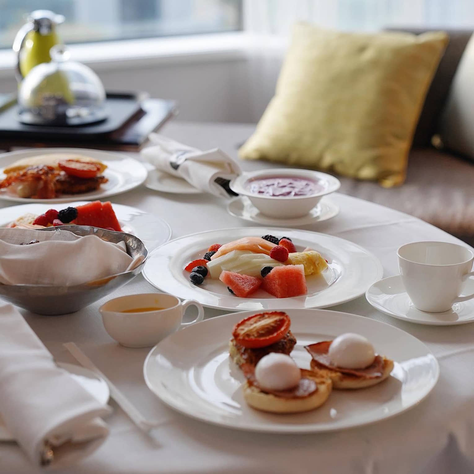 An In-Room Dining breakfast featuring eggs Benedict, fresh fruit, yoghurt and coffee, elegantly arranged on a table by a window with soft pillows in the background.