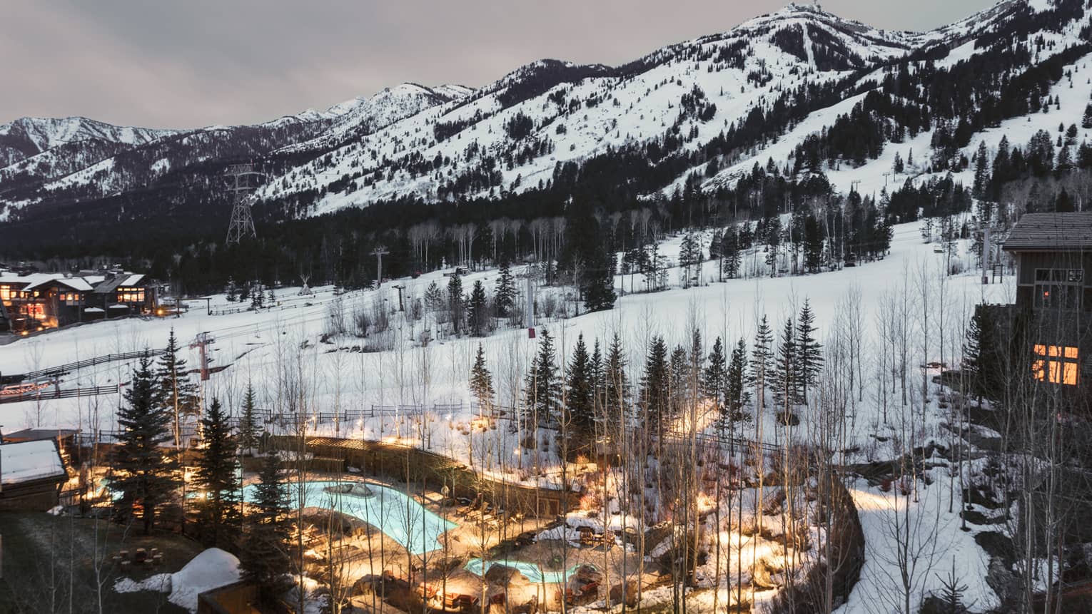 Aerial view of illuminated blue outdoor swimming pool through trees on snowy hill at dusk