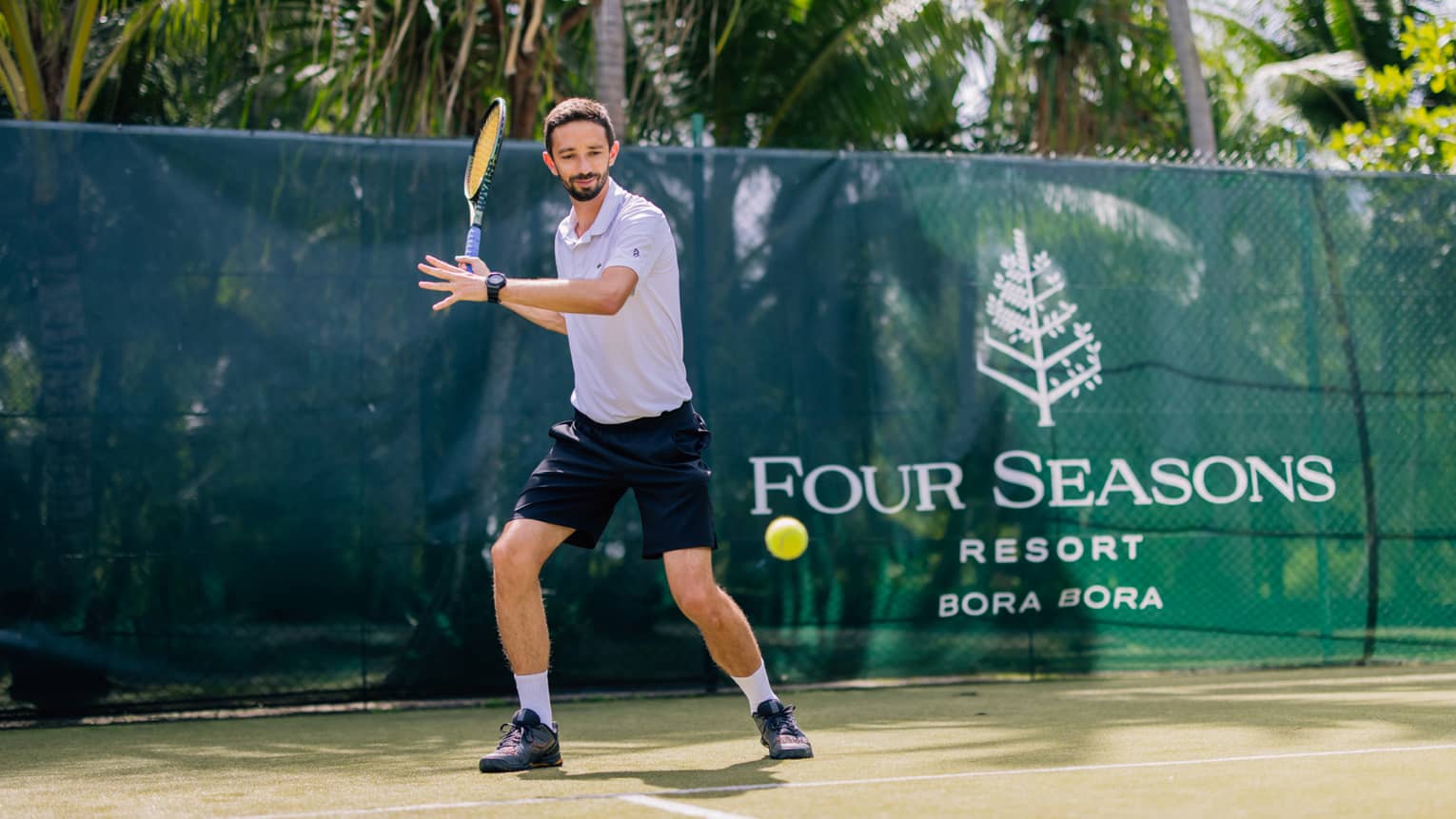 Standing behind the baseline on a court surrounded by palm trees, a tennis player prepares to hit a forehand.