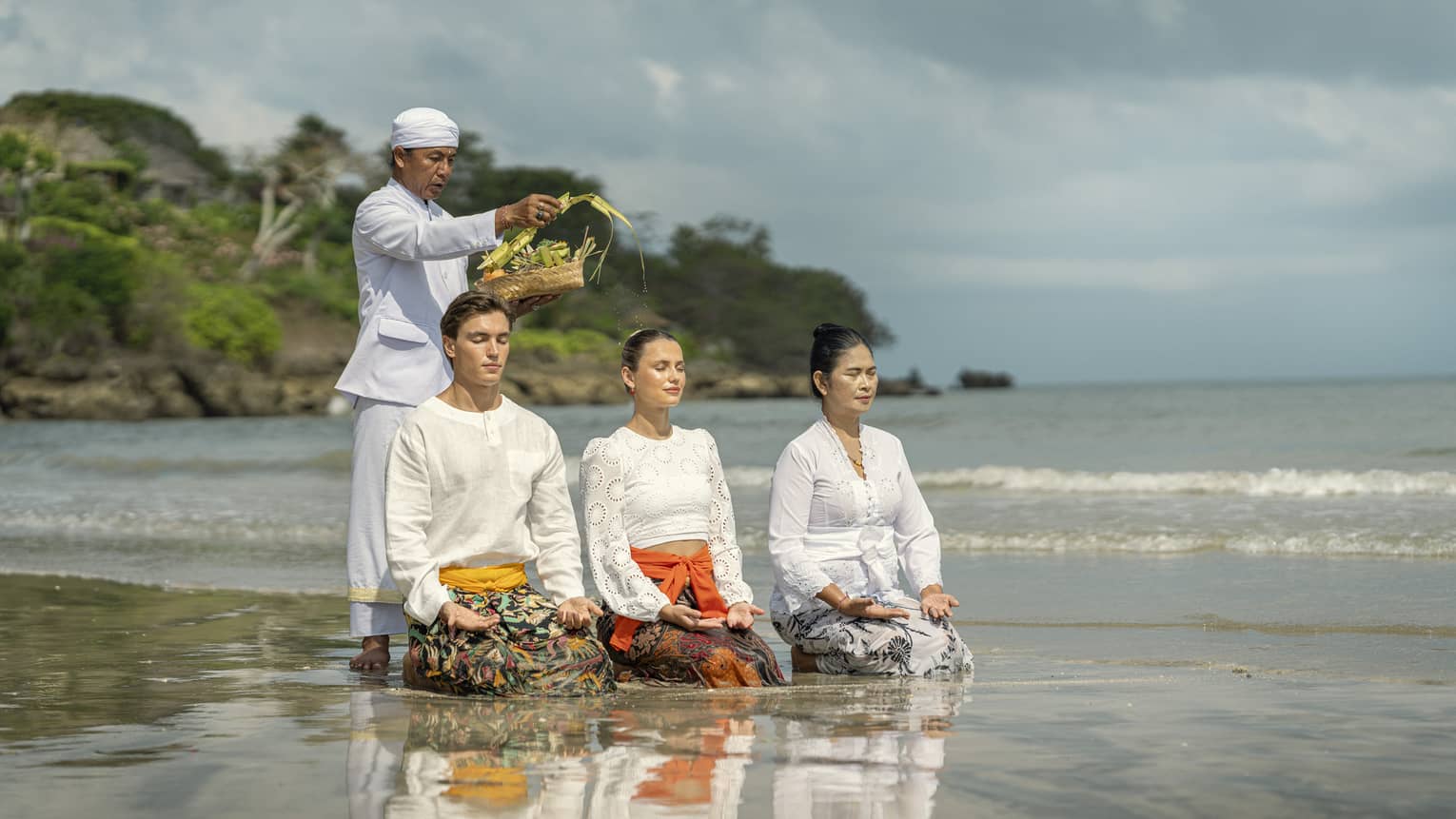 Three adults sit in shallow water with palms up on their knees as another sprinkles water from palm fronds on their heads.