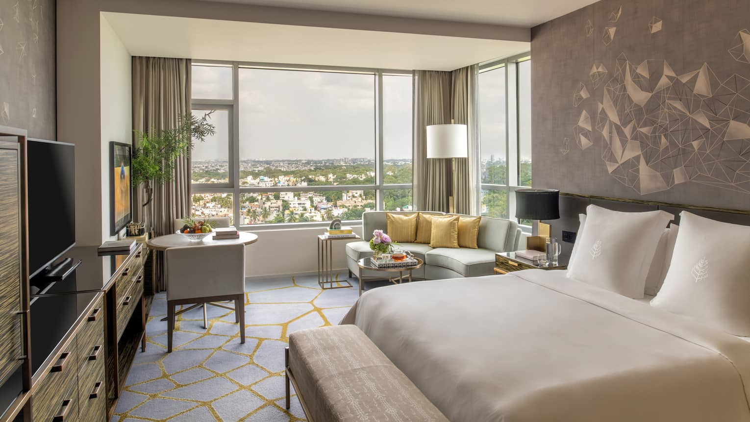 Guest room with king bed and white linens, gold accents, grey corner sofa and windows with city views