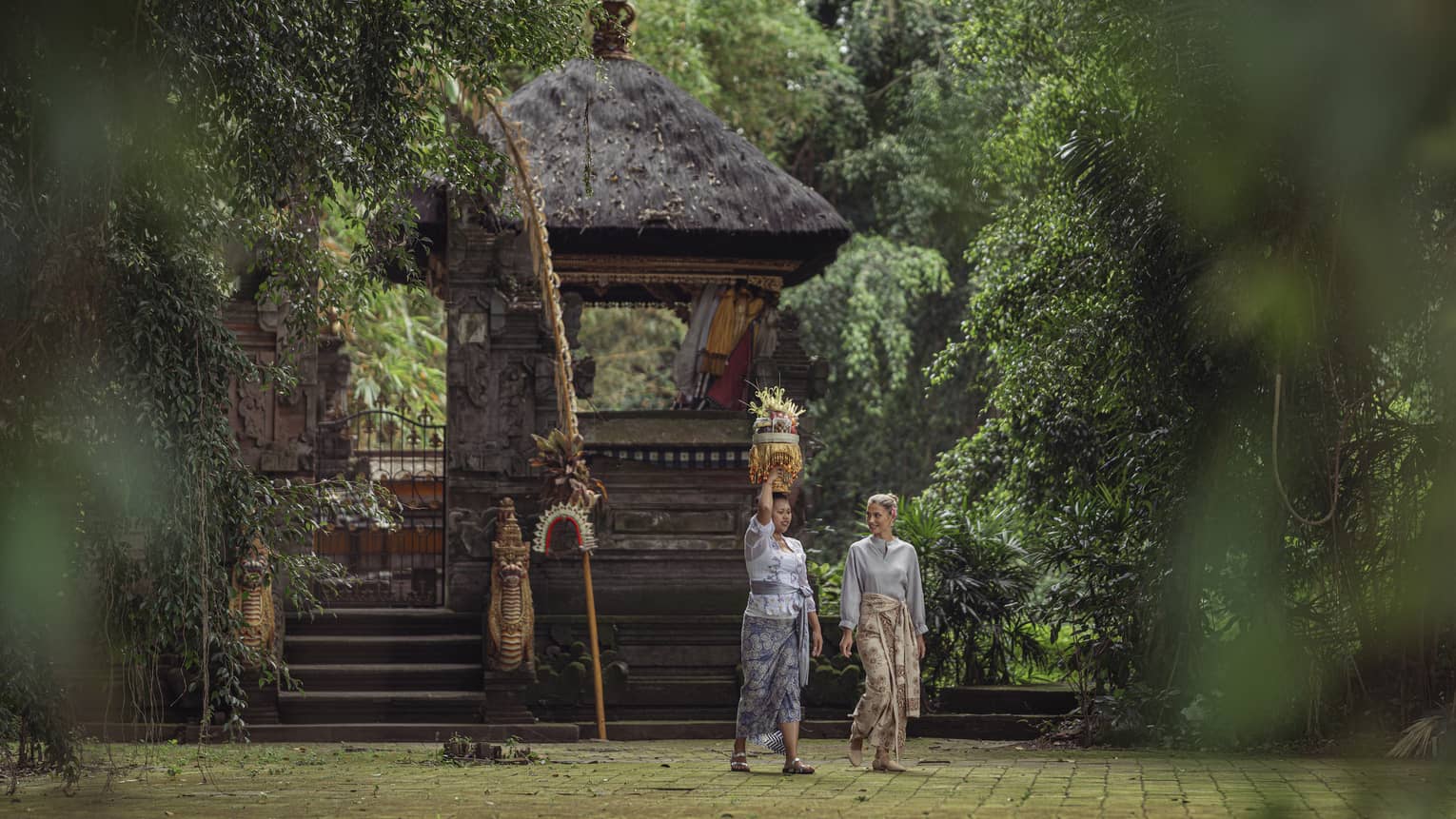 Two women wearing batik skirts walk by a temple in a lush tropical area as one of them carries a golden tray on her head.