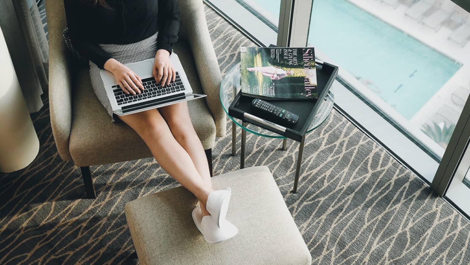 Woman wearing business suit, white slippers works on laptop om legs resting on ottoman