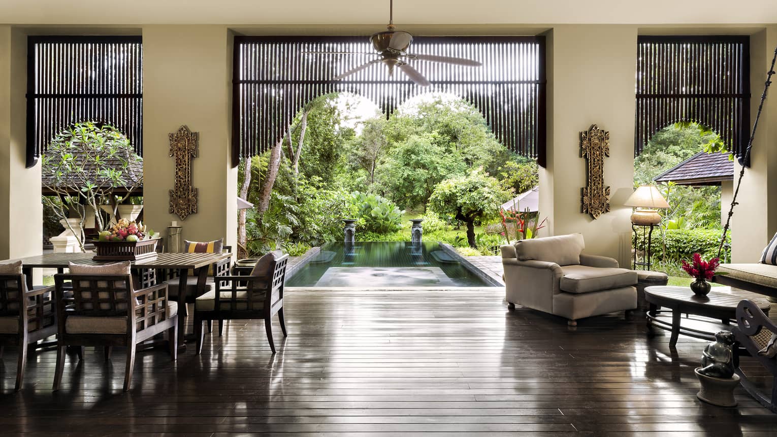 Elegant open-air living space with dark wood floors, carved furniture and dining area overlooking a tranquil pool and lush garden