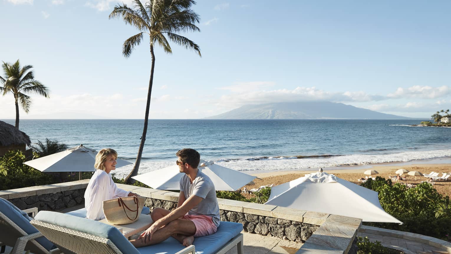 Man in shorts and smiling woman in beach shirt sit on edge of lounge chair, beach and ocean in background