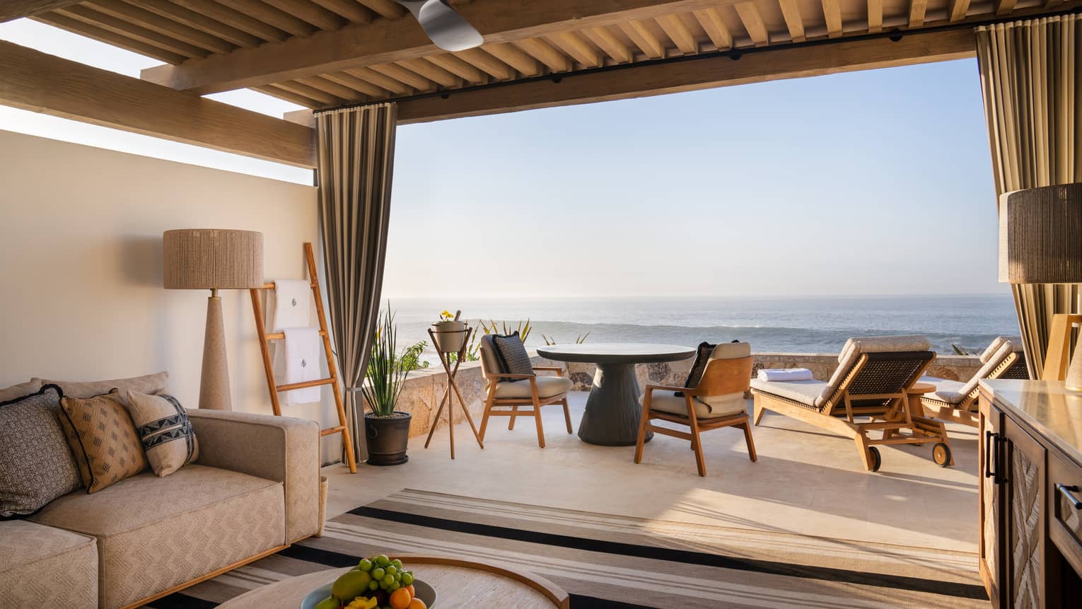 Oceanfront cabana with beige sofa, lamp and round wood coffee table under a roof with a round table and chairs and two lounge chairs out on the open-air patio overlooking the ocean