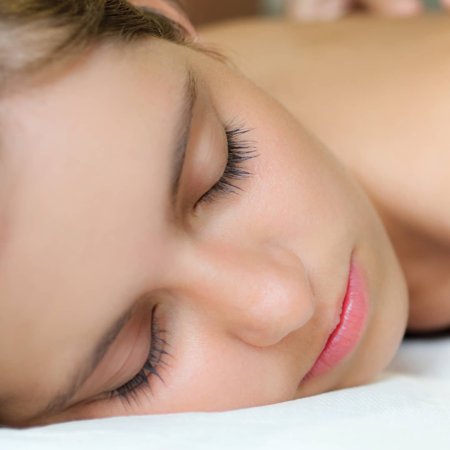 Woman with eyes closed rests on spa treatment table while massage therapist places hands on her bare back