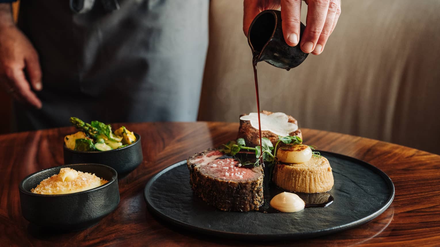 A waiter's hands pour sauce onto a slice of braised short rib served on a black plate next to two small black side bowls