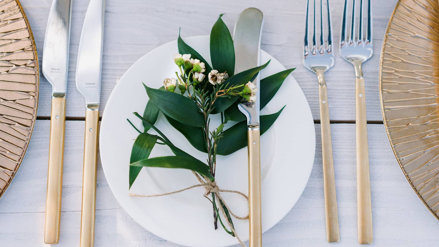 A bohemian table setting of gold chargers, modern long gold and silver flatware, white porcelain plates and a sprig of greenery
