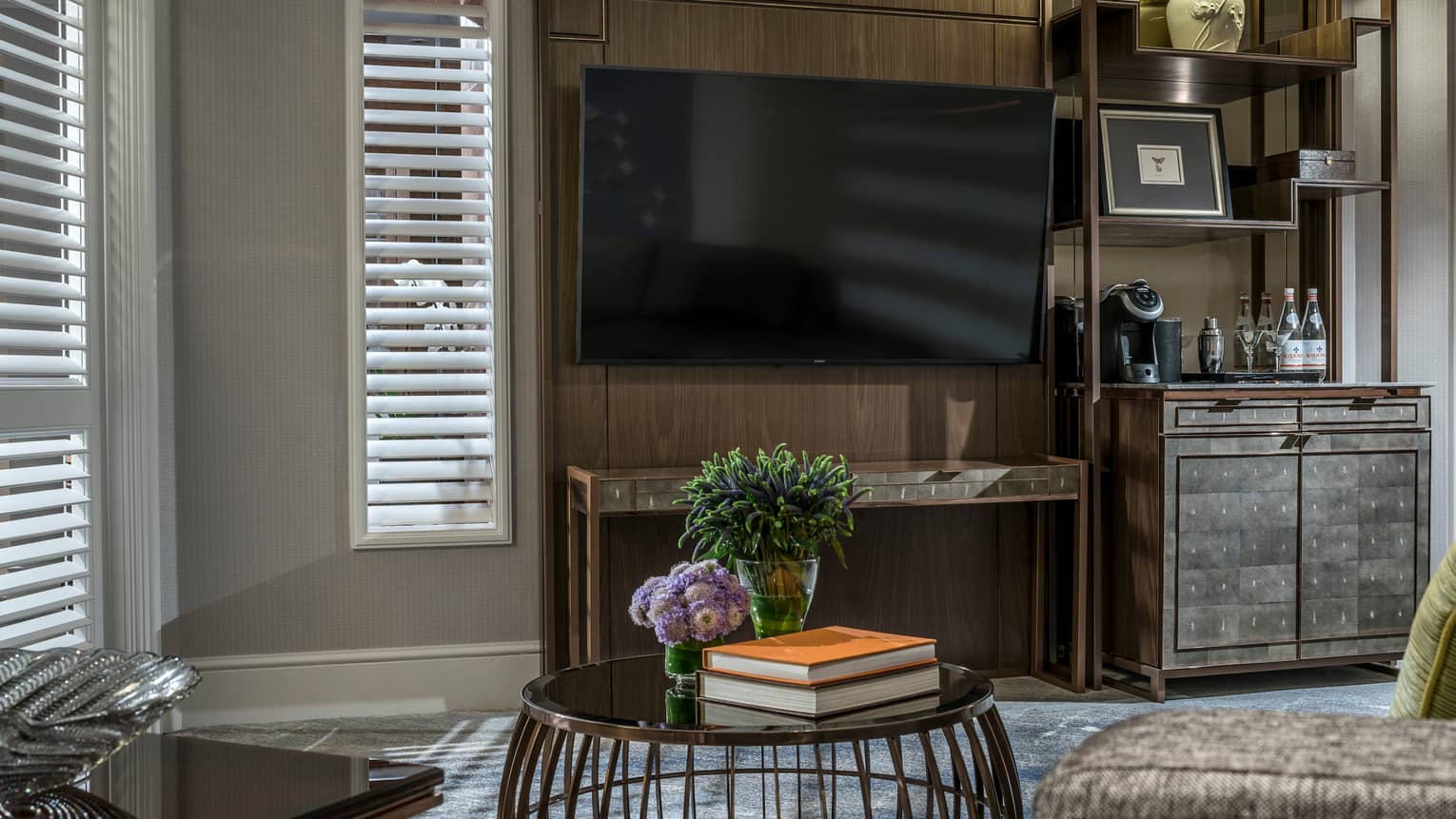 A living area featuring a large wall-mounted TV, sleek wooden cabinet with a coffee machine, and a round table holding flowers and books. Shuttered windows allow soft light to filter in.