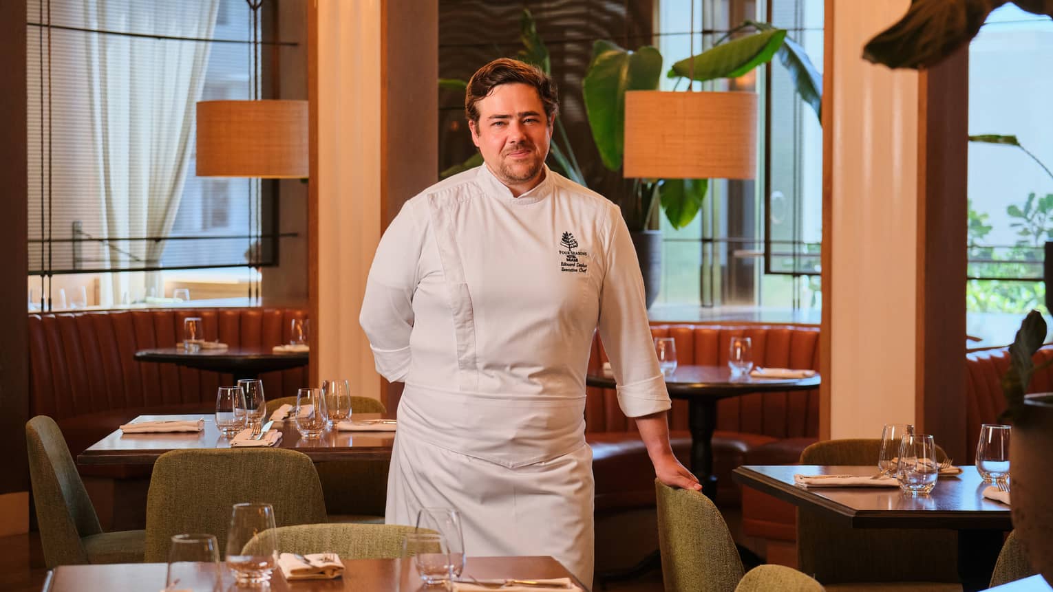 A chef stands in a restaurant space with tables, chairs, soft lighting and large windows.