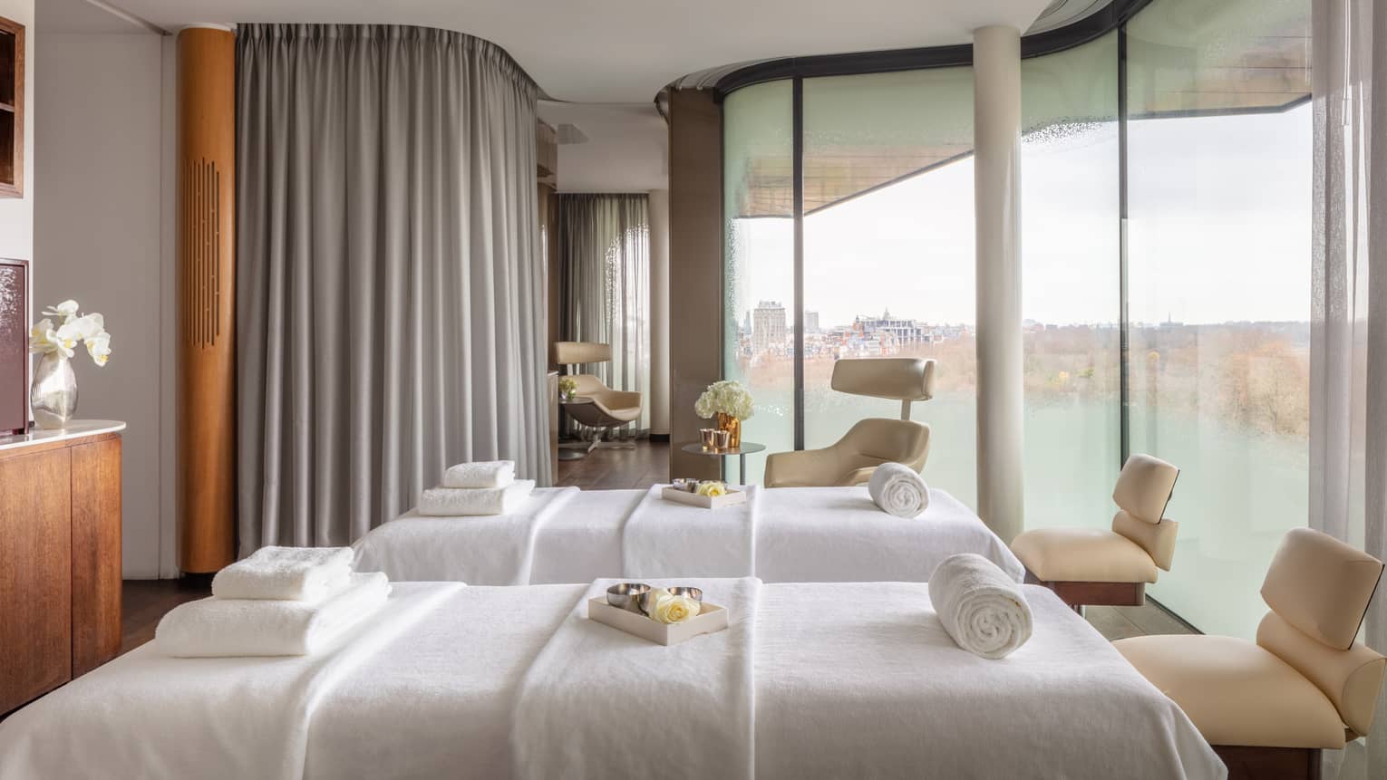 Spa couples treatment room with two white beds, chairs at ends, tall windows with city views