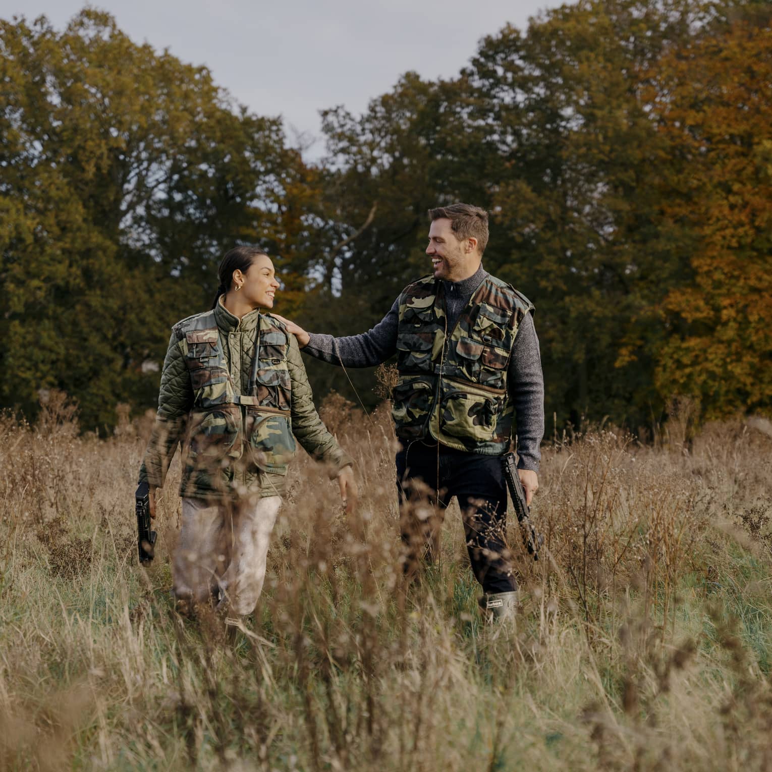 Two people wearing camouflage vests stand in a grassy field with trees in the background.