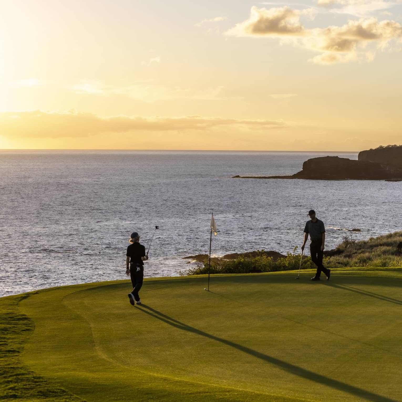 The setting sun silhouettes a pair of golfers enjoying a game of golf. The golf course is on top of a cliff beside the ocean.