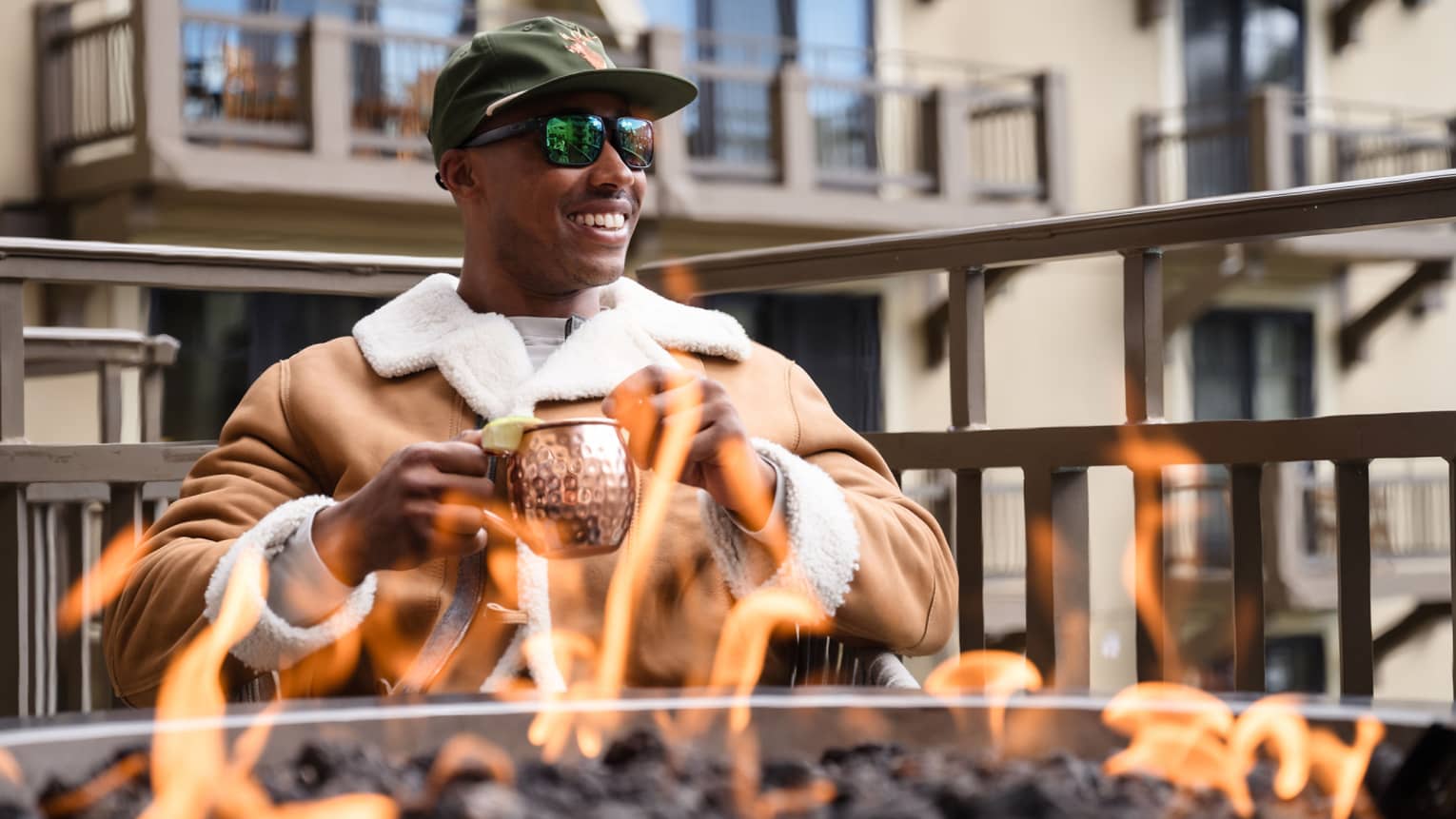 Guest wearing a jacket and holding a mug while sitting by a fire pit.