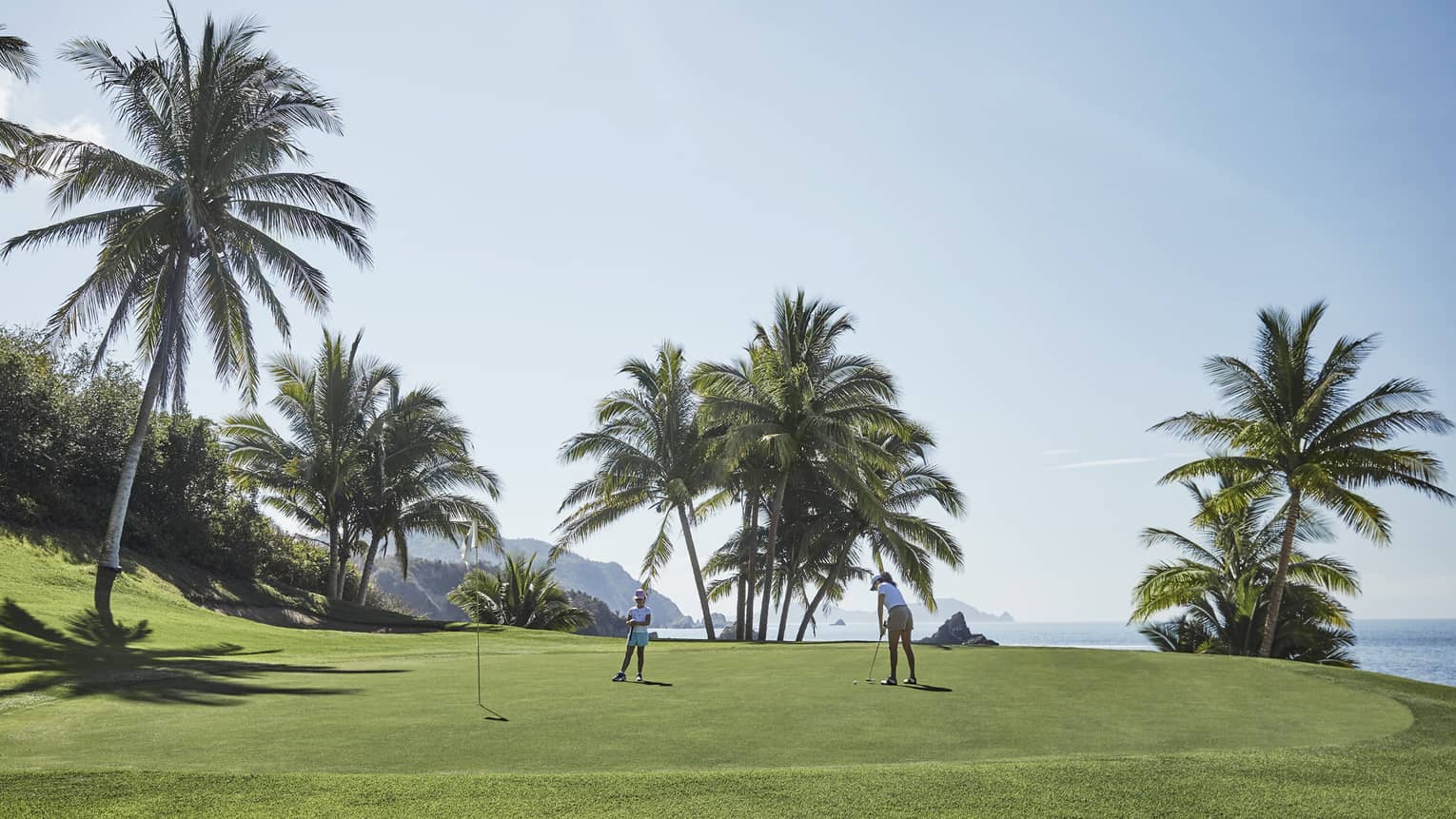 A golfer about to tap a ball toward a nearby hole as a child looks on, on a pristine green bordered by ocean and palm trees.