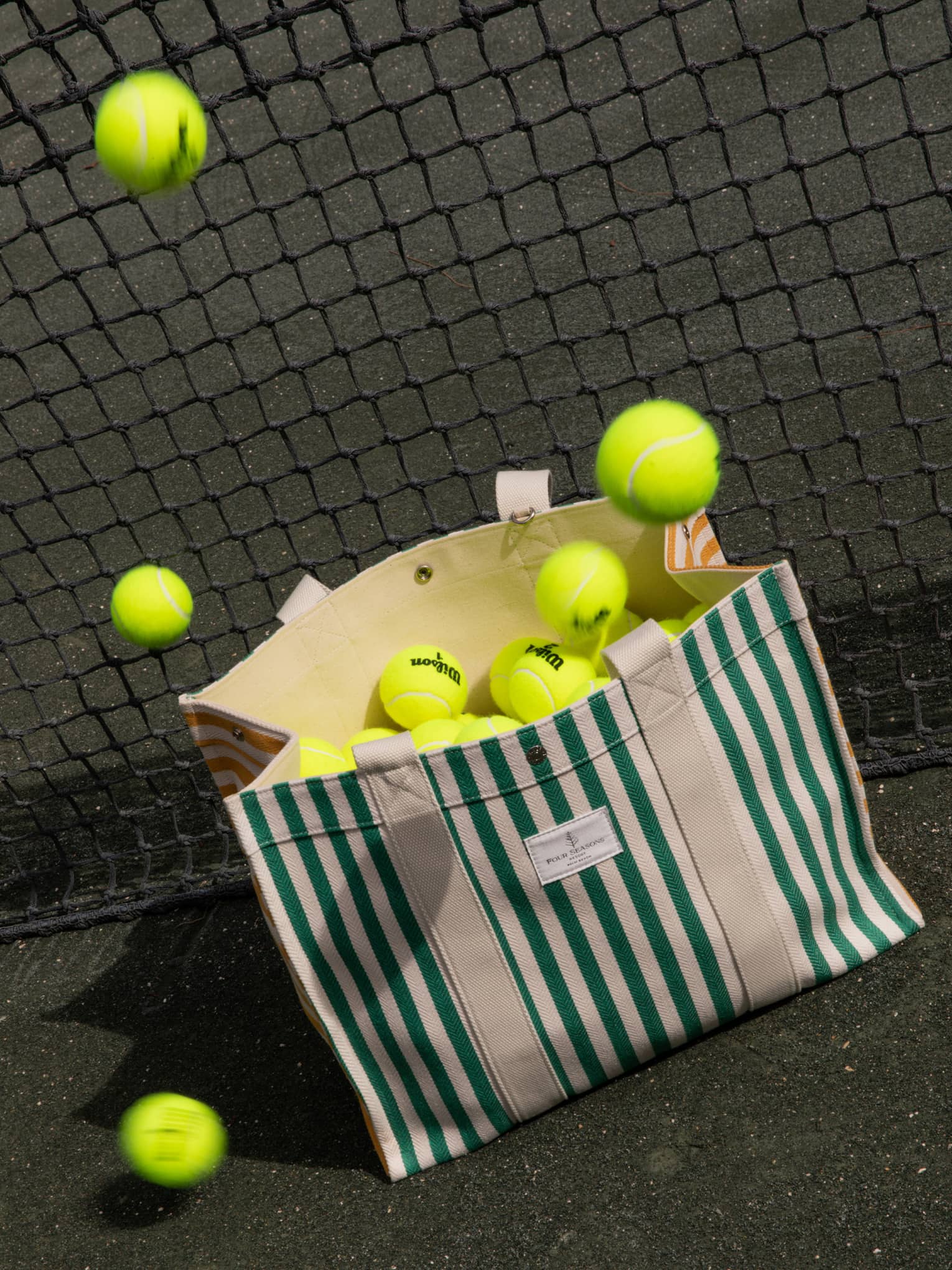 Green-and-white striped canvas bag filled with tennis balls leans against a net