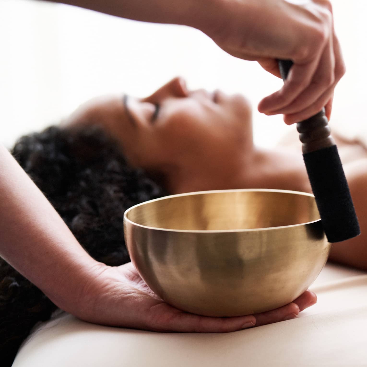 Woman with eyes closed lies on massage table in as vibrational sound therapy is performed with a Tibetan singing bowl