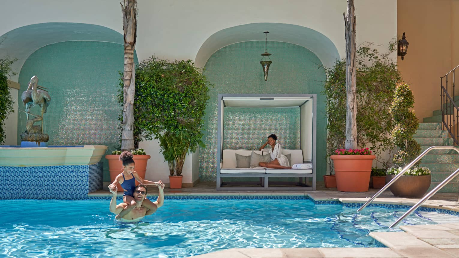 A man and young child swimming in an outdoor pool, while a woman looks on from a cabana