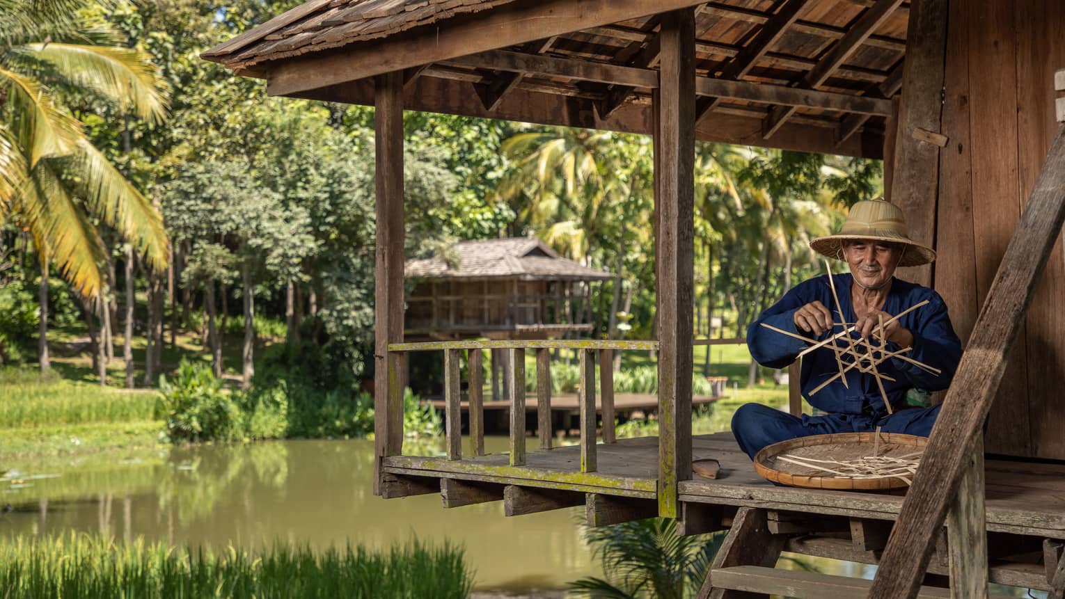 Man weaves a traditional Thai basket on the porch of a riverside pagoda