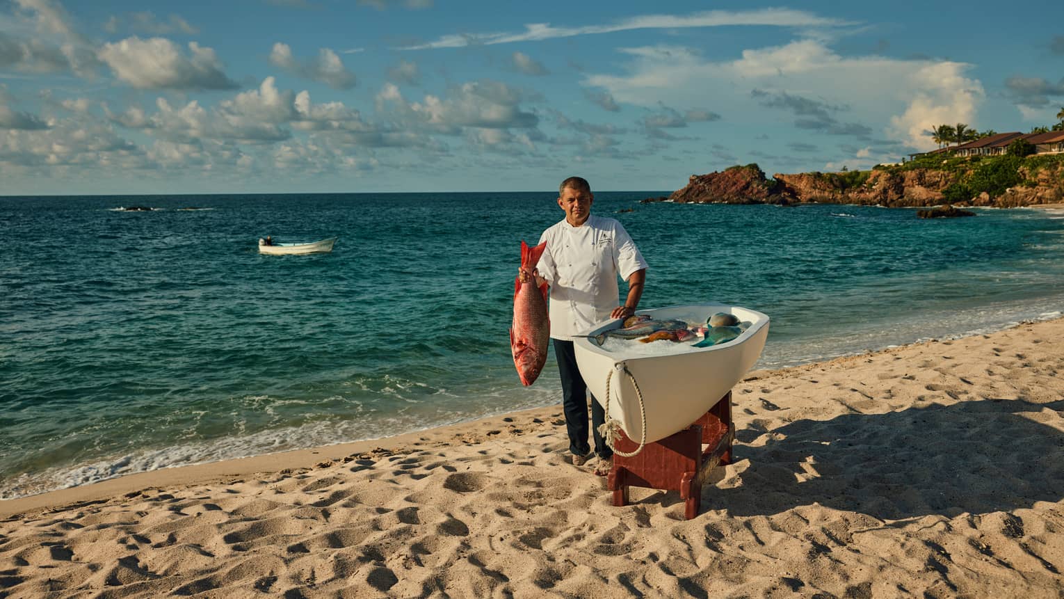 Chef in white chef’s coat, standing on beach beside a small boat, holding a freshly caught fish