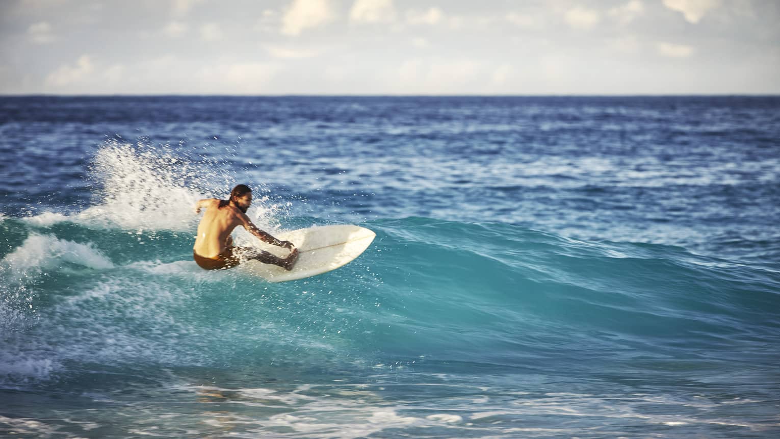 On an expanse of water, a surfer leans back on his board and surfs the face of a wave as white water splashes up behind him.