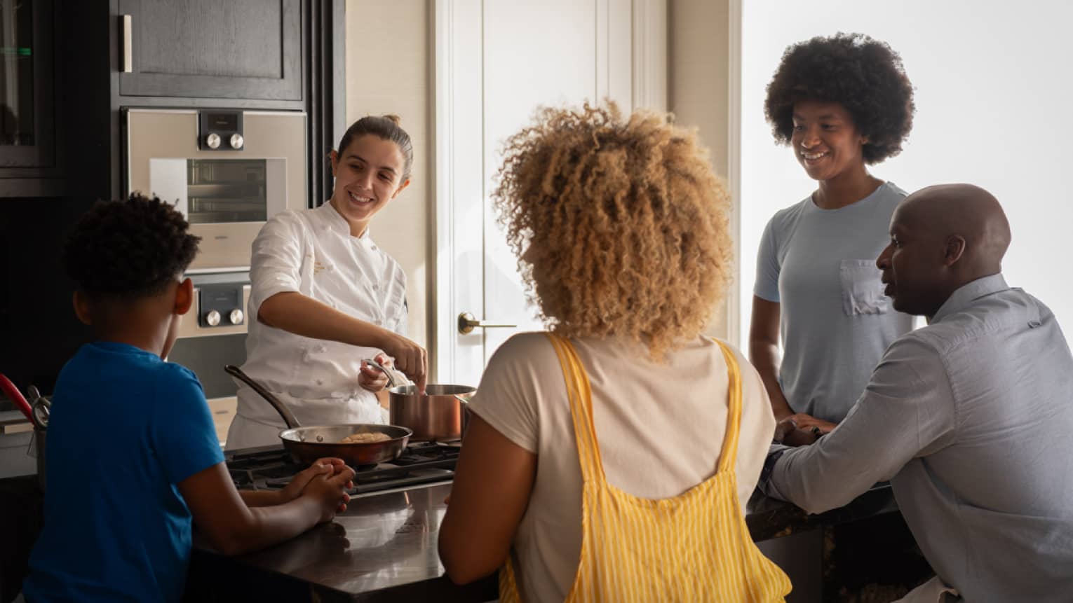 A family of four gathers around a kitchen-island cooktop watching a smiling chef prepare food in copper cookware.