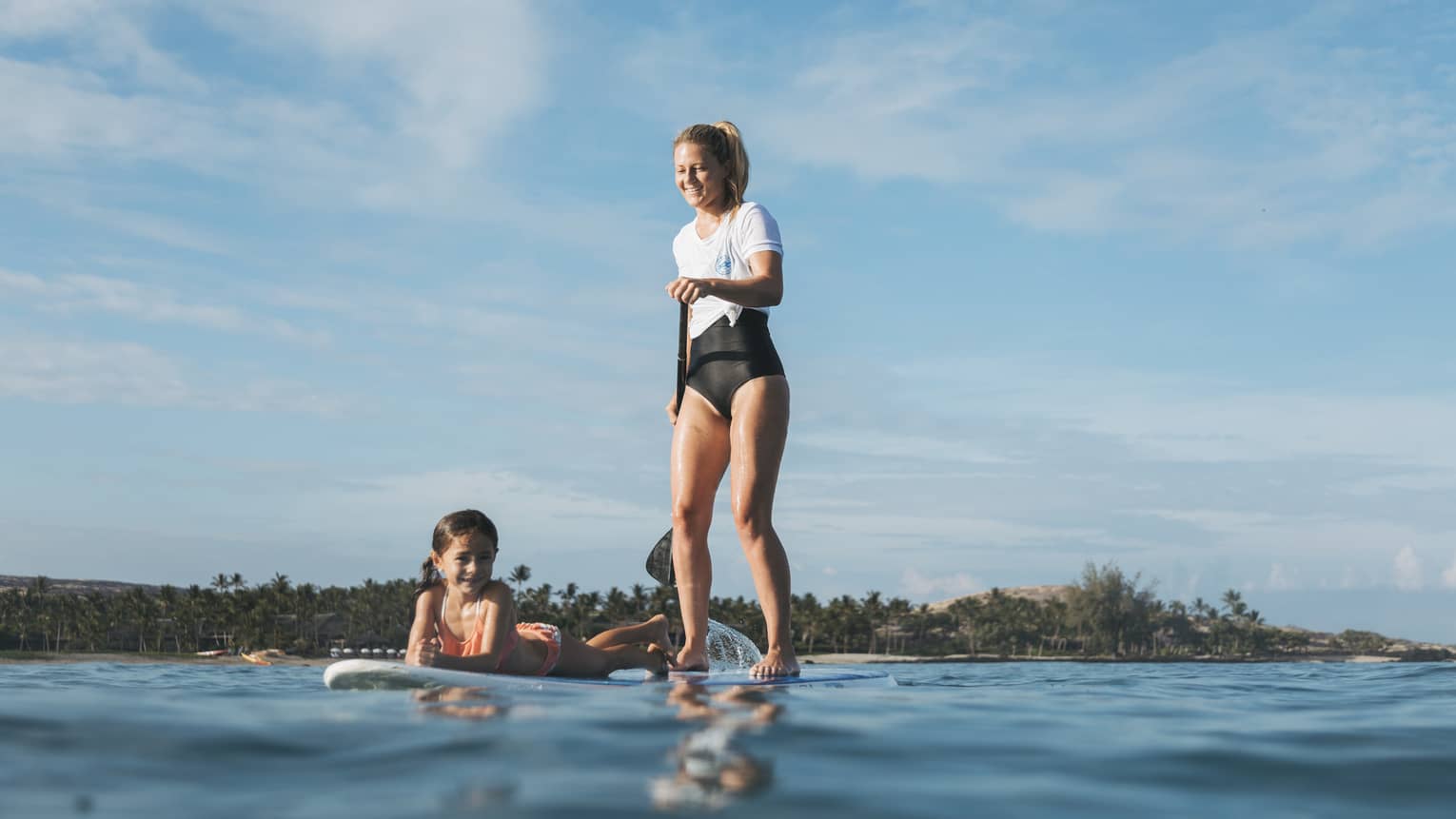 A smiling parent and child enjoying a shared paddleboard ride in open water with the shoreline in the distance.