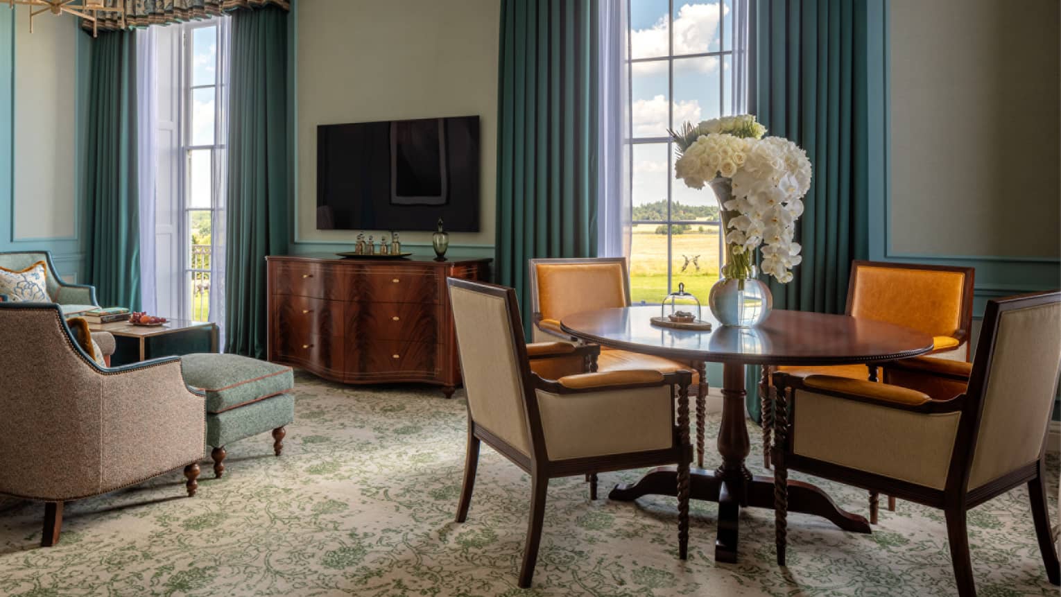 Guest room with round wooden table and four dining chairs, cherry sideboard, flat screen TV, blue chaise, twin windows with blue drapes