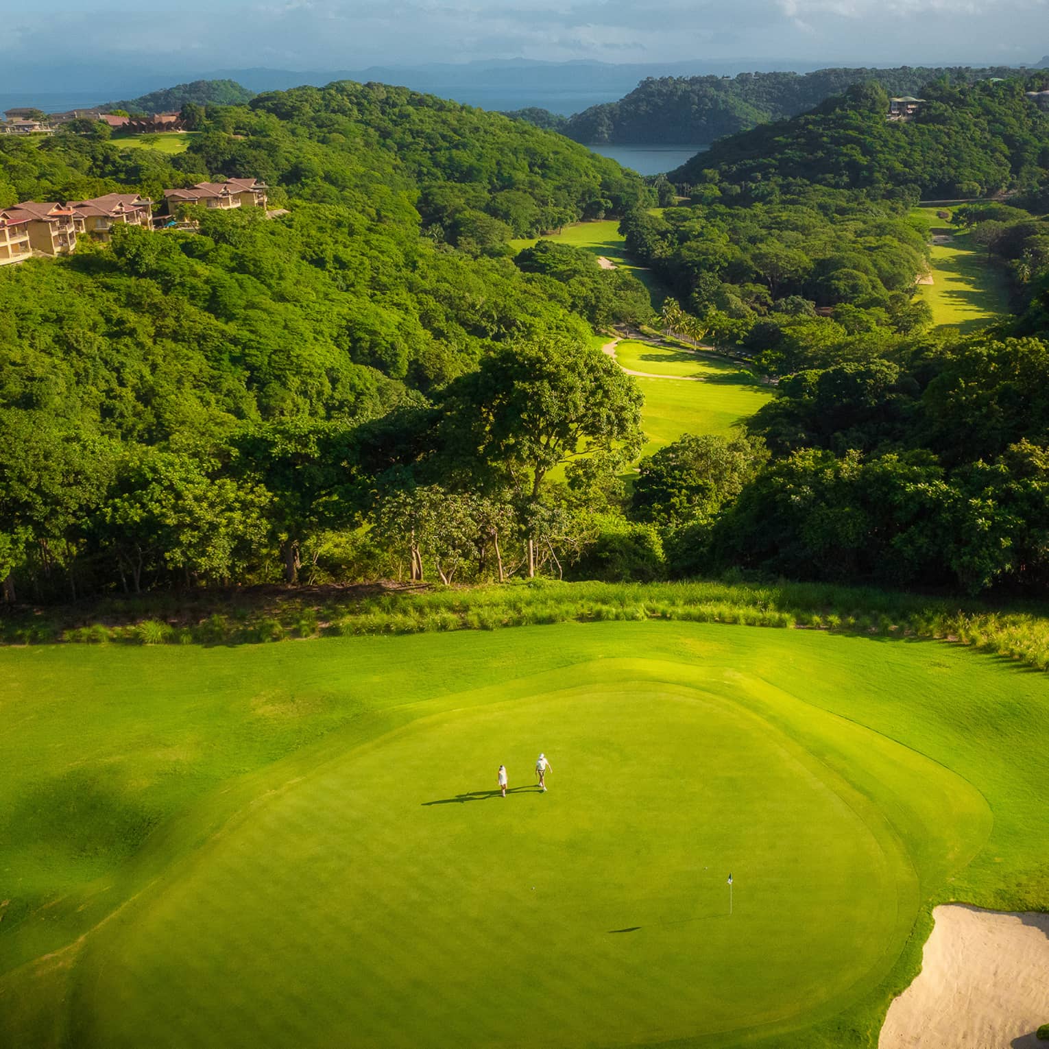 Aerial view of two people standing on a golf green with tree-covered rolling hills behind them leading to the ocean in the distance
