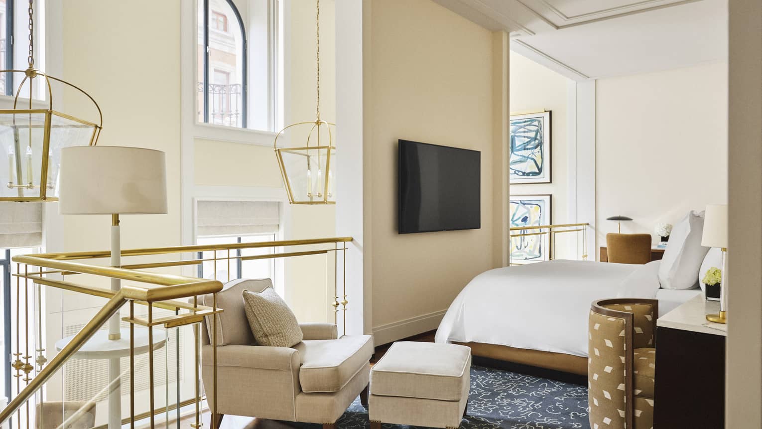 Luxury hotel loft bedroom with a bed, wall-mounted TV, beige armchair and ottoman near a gold-railed balcony, hanging lantern-style lights and abstract wall art