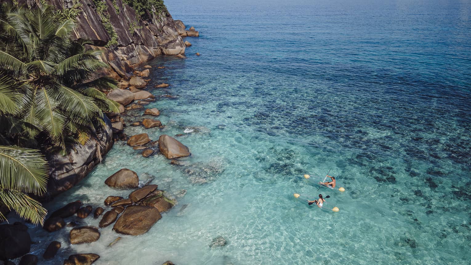 Kayakers in transparent kayaks paddle towards a rocky cliff covered in greenery, while surrounded by crystal-clear water.