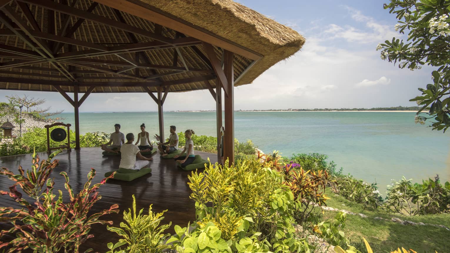 Five people sit cross-legged meditating on green cushions under thatched-roof gazebo by ocean