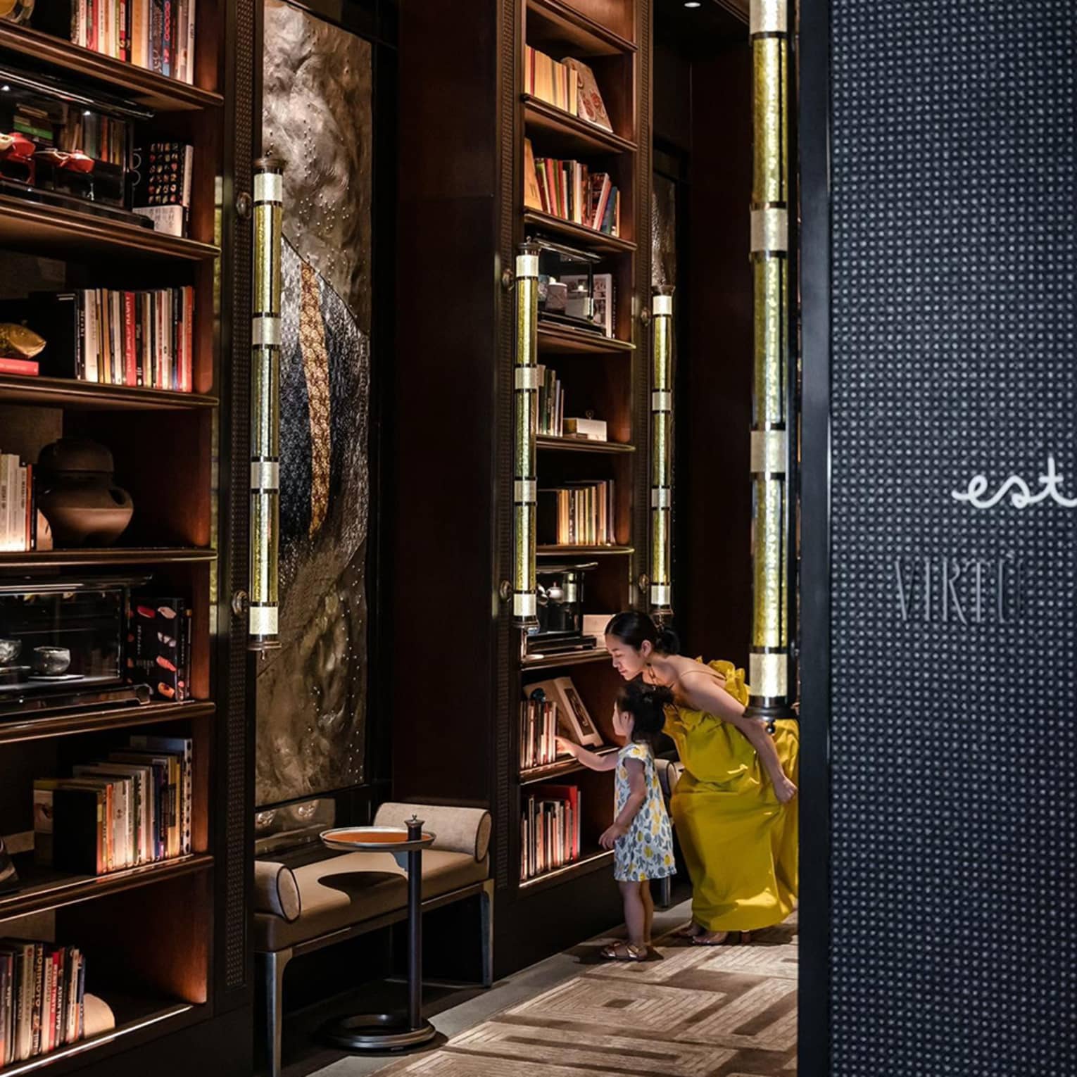 Adult and child exploring a dimly lit library with tall bookshelves in an elegant setting