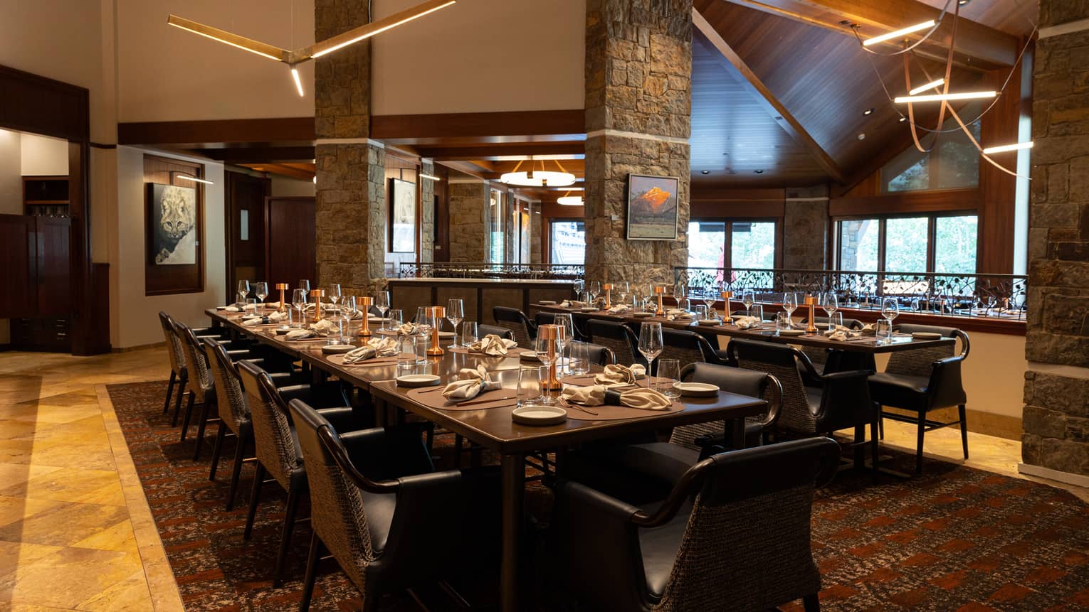 A warm interior dining space with windows on one wall, wood floors, long dining tables set for a meal, chairs, and large three-bladed ceiling fans suspended from the high ceiling