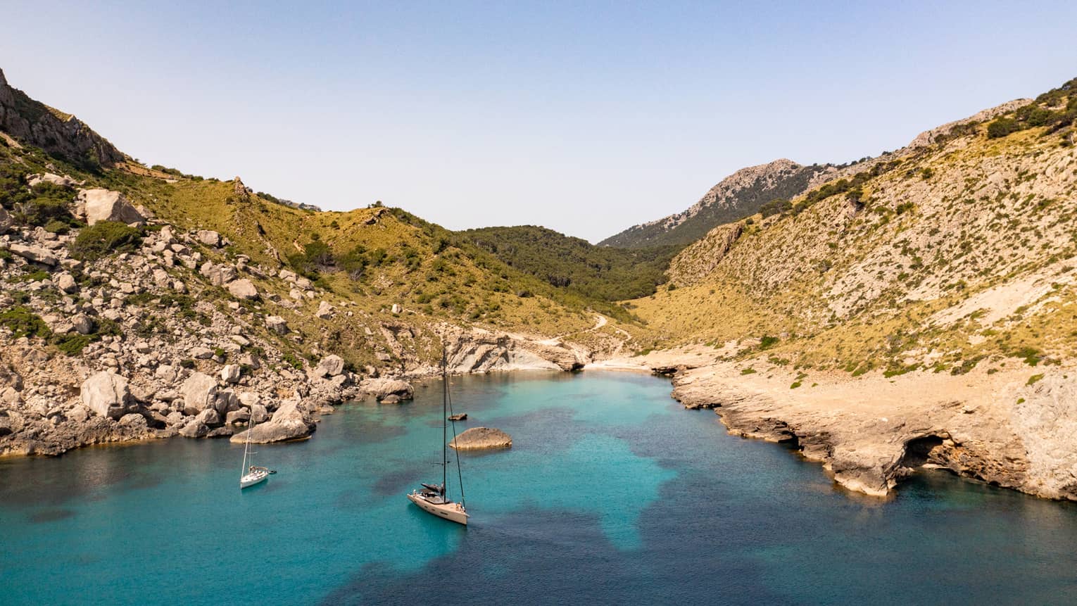 Aerial view of two boats in variegated azure water under a clear blue sky in a cove framed by rocky hills dotted with shrubs.
