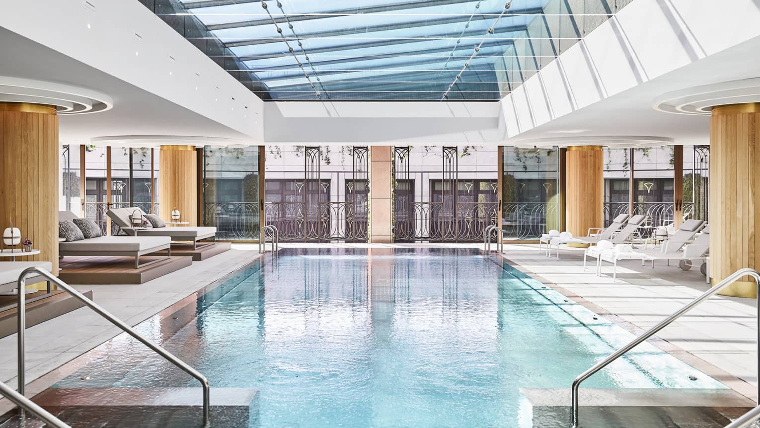 Luxury hotel indoor pool area with glass ceiling, metal railing accents and lounge chairs, surrounded by large windows and architectural detailing