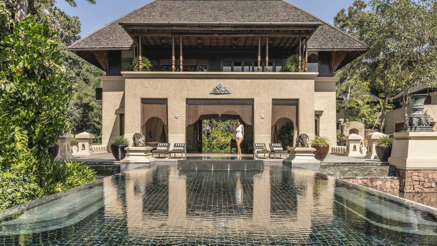 Person walking along the edge of a large reflective pool in front of a grand villa with traditional architecture surrounded by trees and outdoor seating