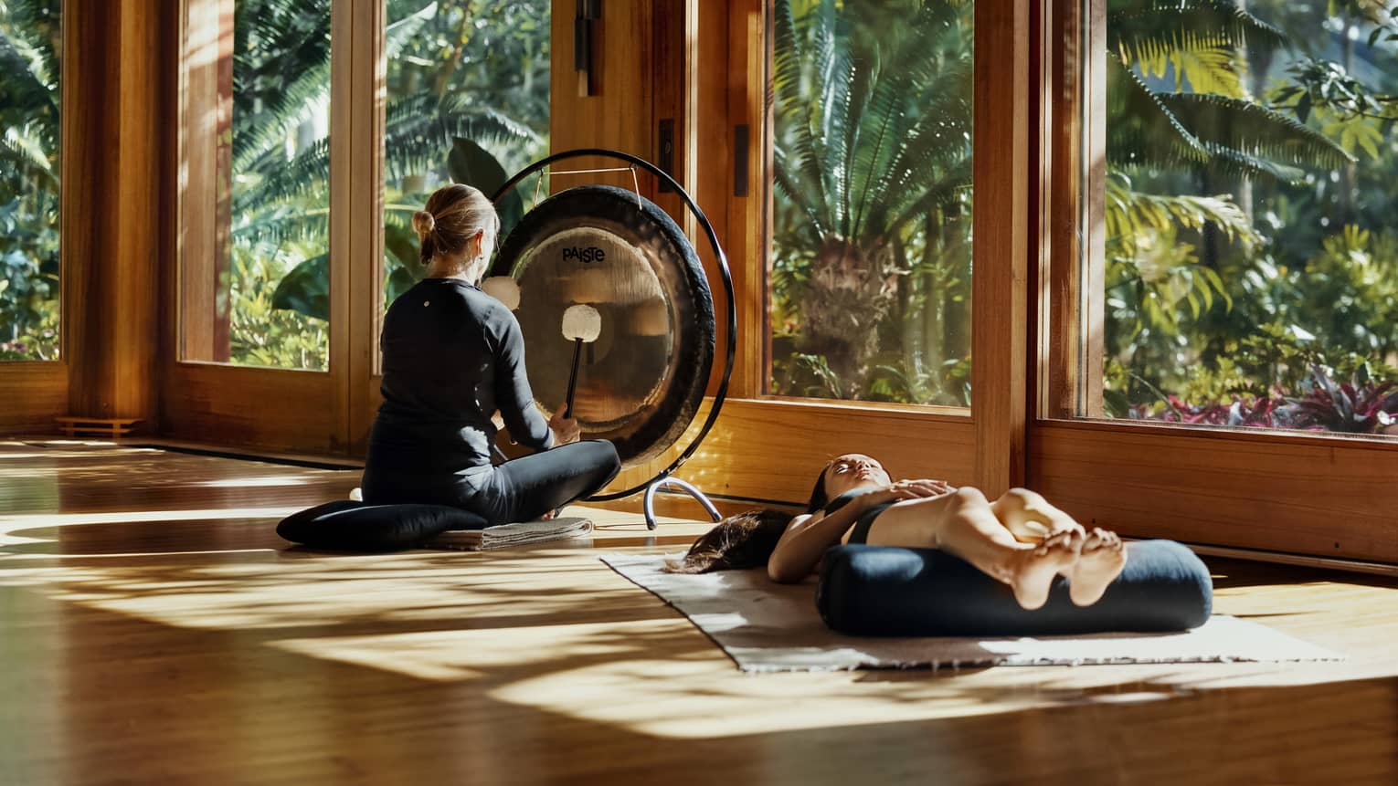 During a sound bath, a meditator lays on a mat, legs resting on a bolster, as a practitioner plays a gong with two mallets.