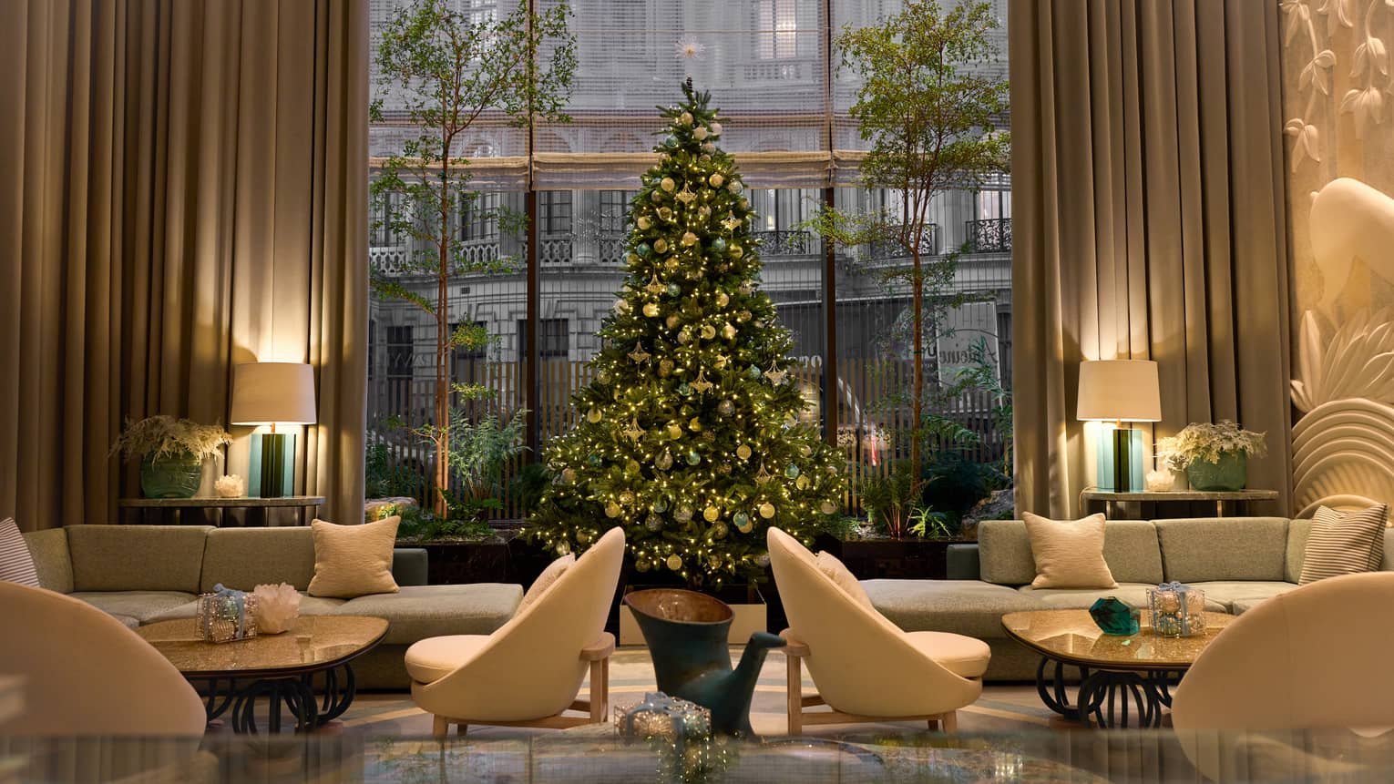 A Christmas tree decorated with gold and teal ornaments stands before a large window in a neutral-coloured seating area.