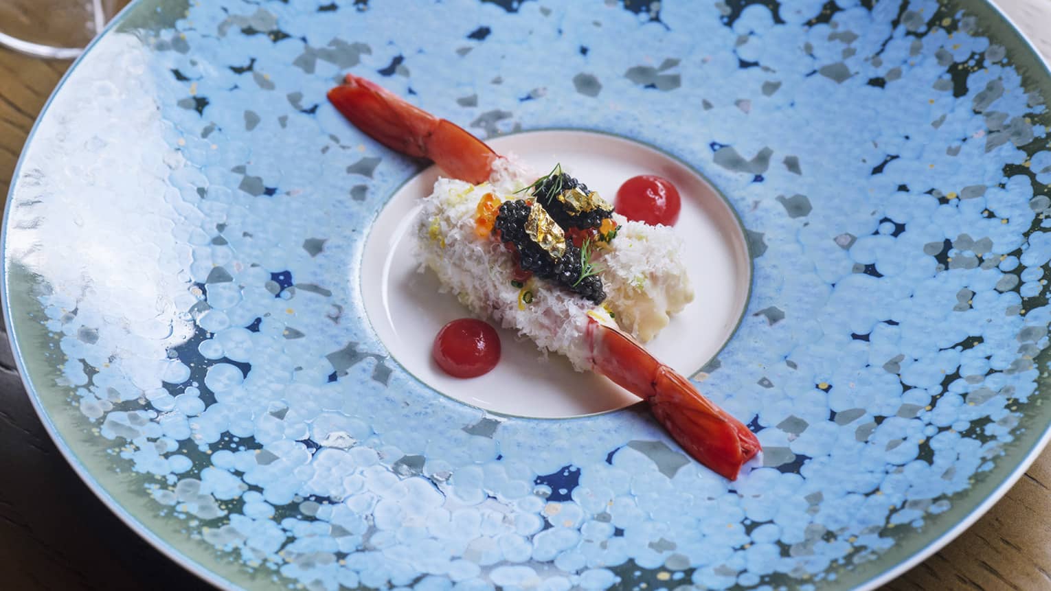 Two elegantly plated prawns topped with garnishes and flanked by cherry tomato halves in centre of blue speckled plate