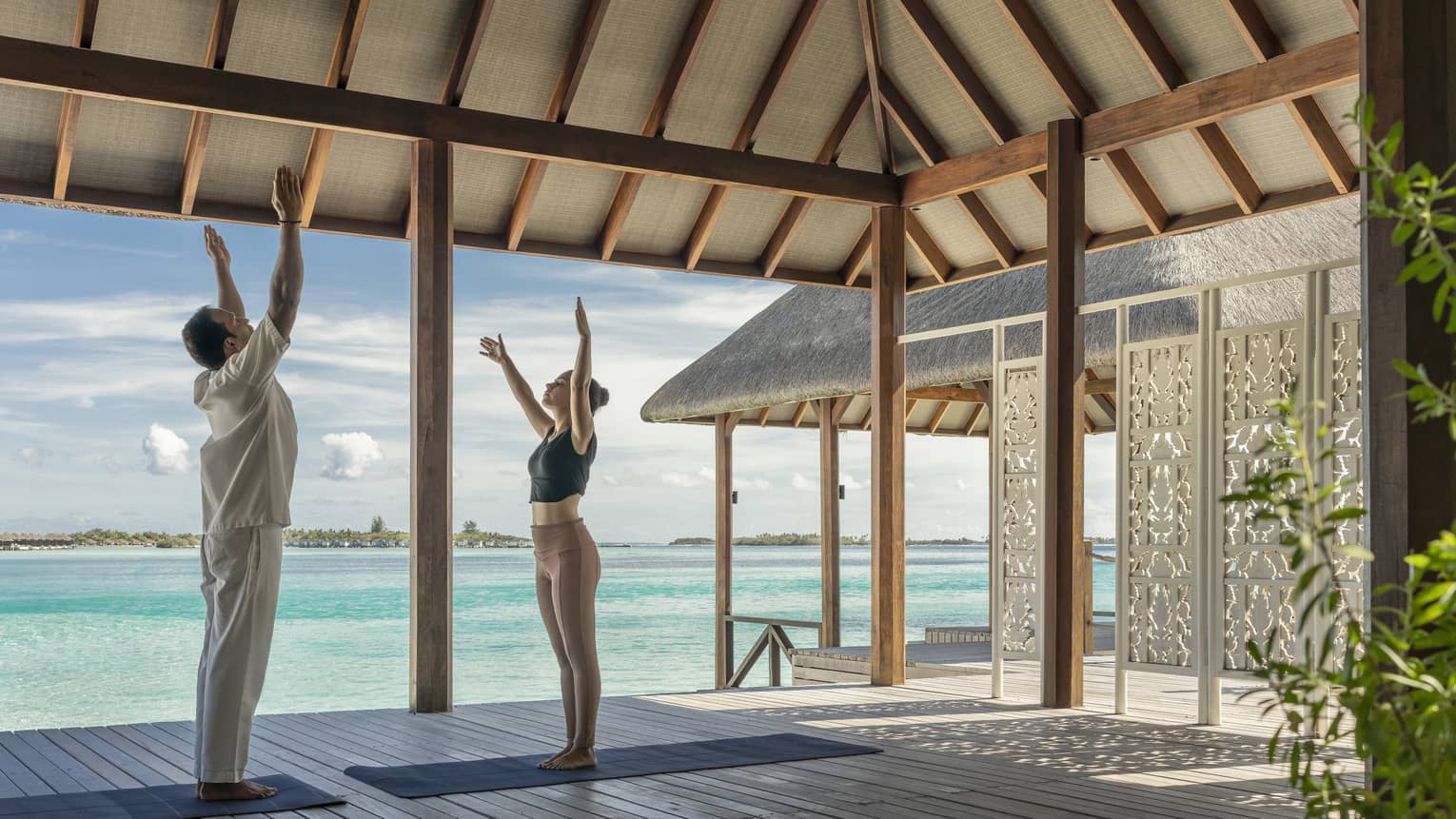 Two people practising yoga on a shaded deck overlooking turquoise ocean water at a tropical island retreat