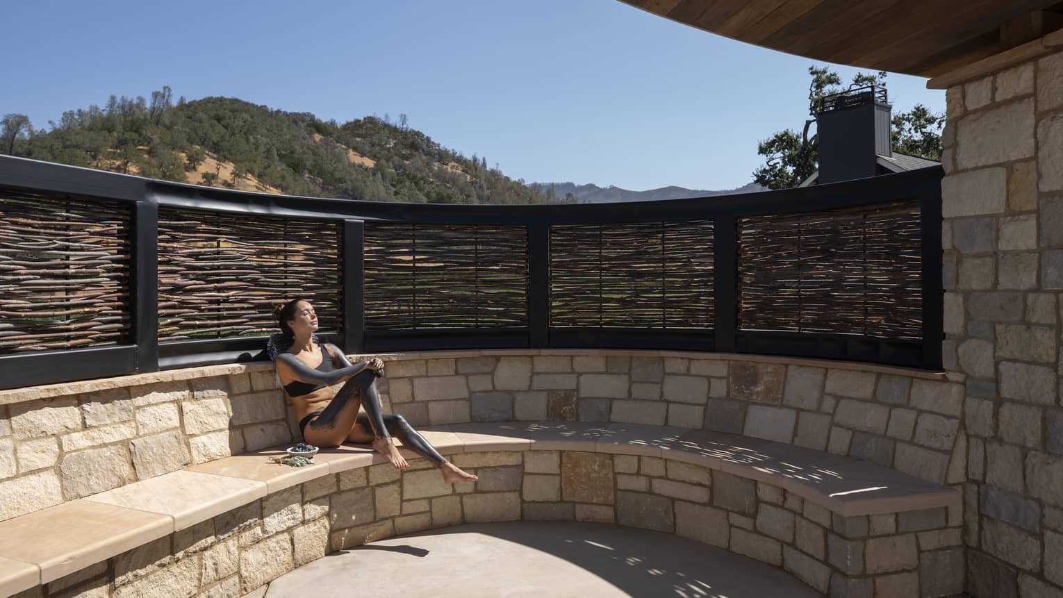 Woman slathered in mud sits on stone bench sun tanning during spa treatment