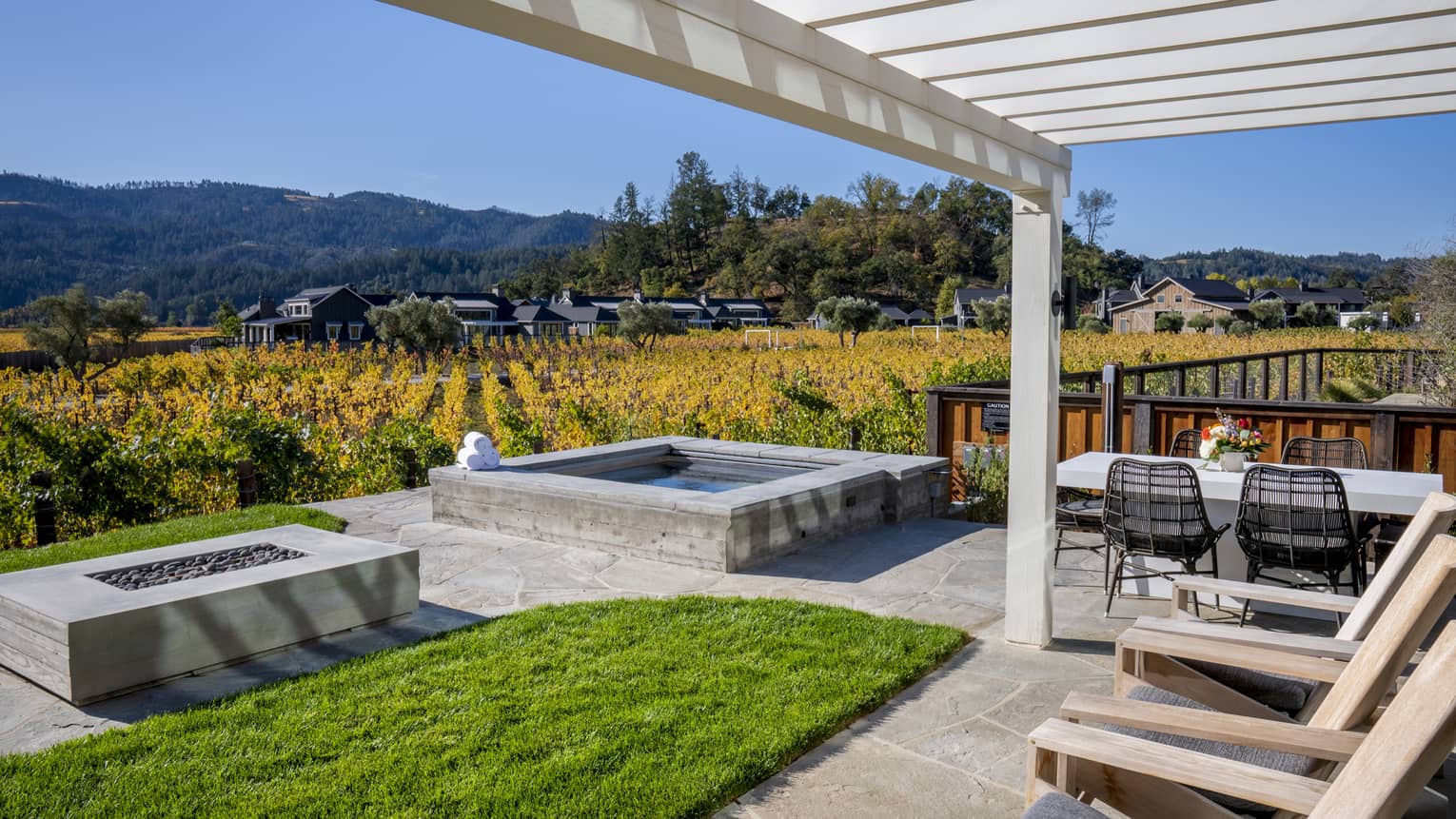 Outdoor villa patio with hot tub, fire pit, lounge chairs and dining table, overlooking vineyards and hillside scenery under a pergola