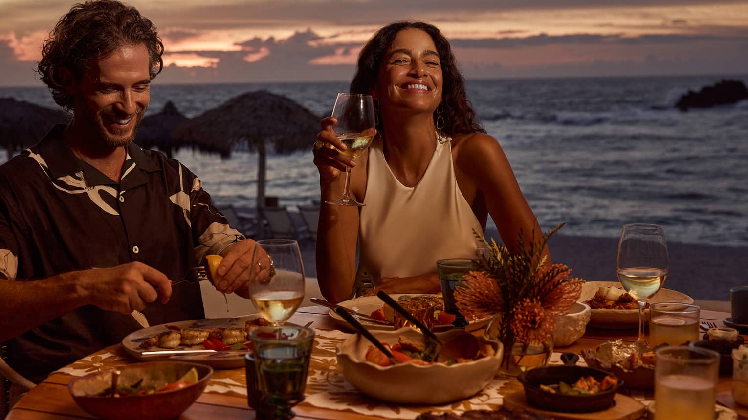 Man and woman laugh while dining at outdoor table filled with food and wine, next to beach and ocean