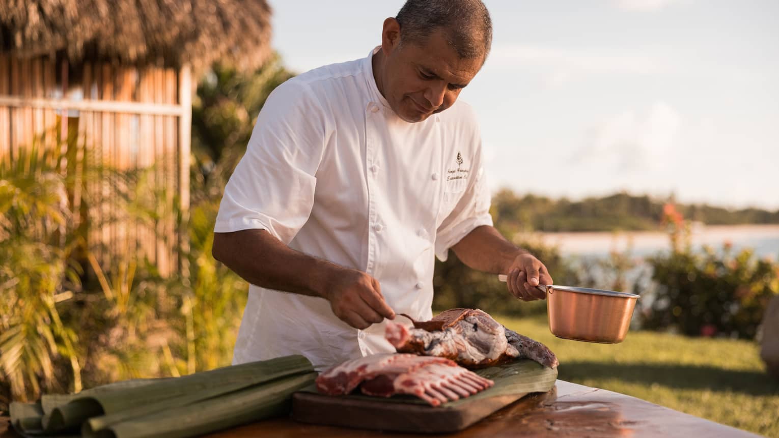 Chef in white chef’s coat, standing at an outdoor table, holding a copper pot in one hand and seasoning meat with the other