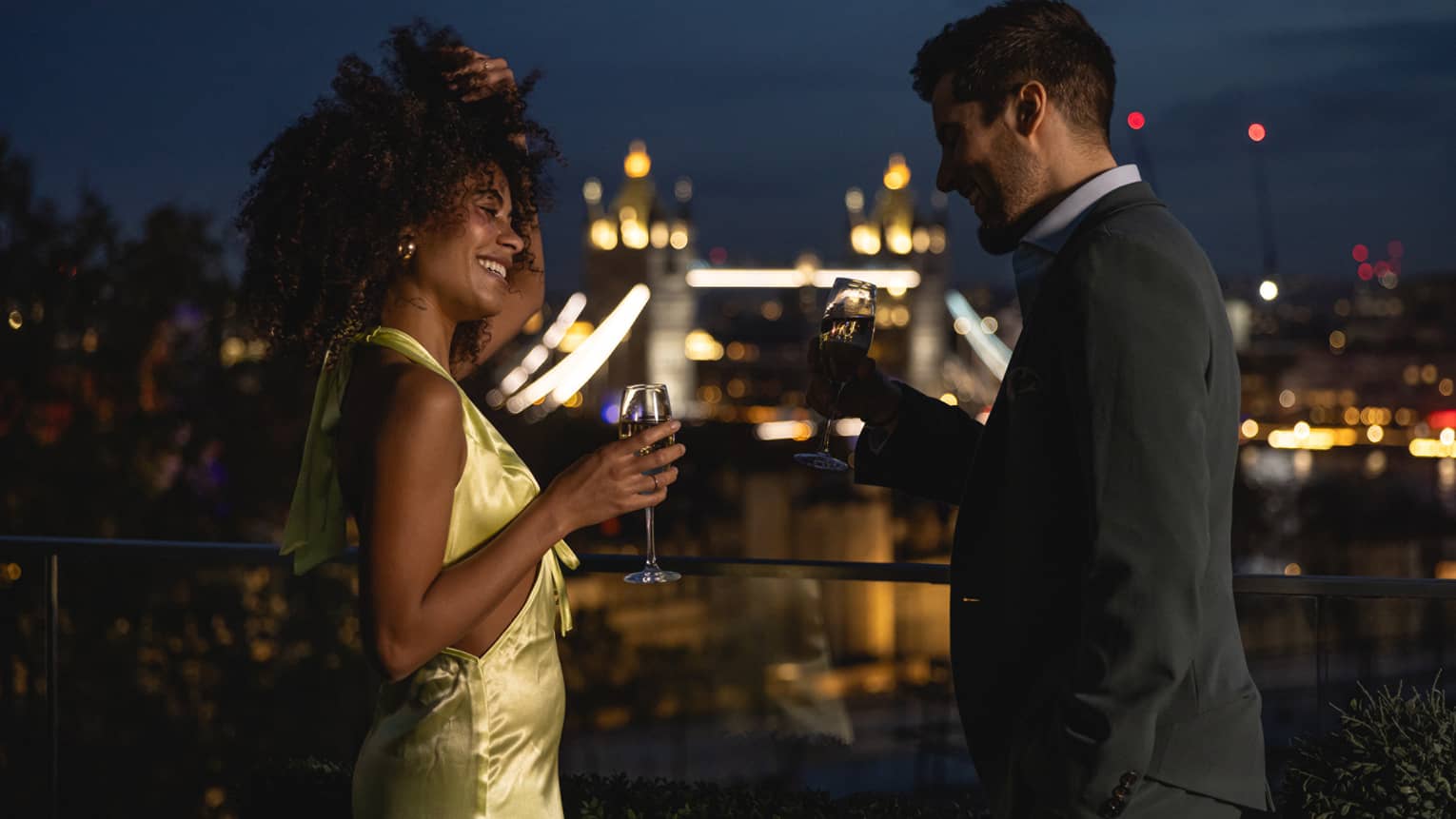 Couple standing on terrace, laughing with champagne in hand over London views at night