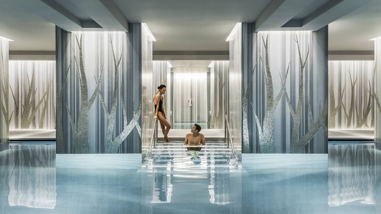 Woman in black swimsuit enters serenity pool while man sits on steps between blue and silver columns