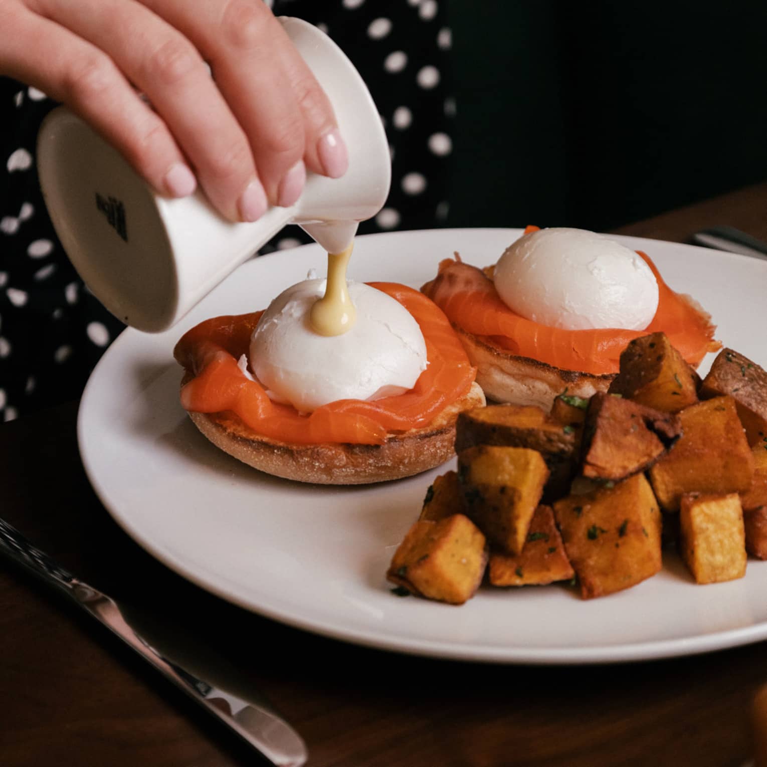 Sauce being poured over two poached eggs with smoked salmon on English muffins, served alongside roasted potatoes.