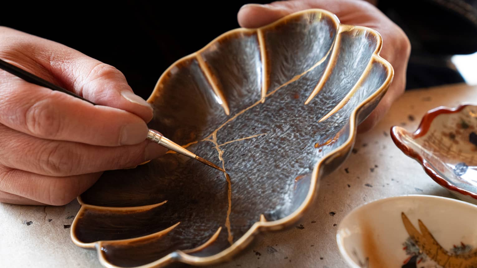 A person using the art of Kintsugi, by using gold paint to paint over the cracks of a once broken black flower shaped plate.